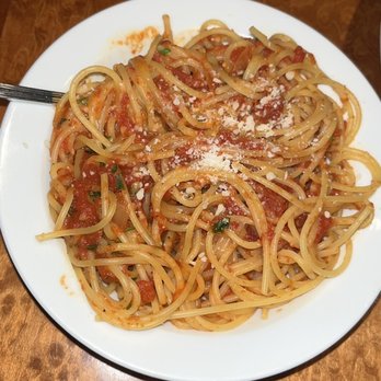 Cafe Amici - Spaghetti with marinara - A round white plate contains cooked spaghetti pasta coated with marinara sauce, garnished with grated cheese and small green herbs. The plate is placed on a wooden table, and a fork is partially visible on the left side of the plate. The background shows a wooden surface, and part of a bottle or container is visible at the bottom right corner. - Italian Near Me - Hamden, Connecticut