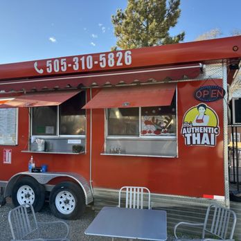 Bo’s Authentic Thai - A red Thai food truck with a phone number displayed on the awning, featuring two serving windows with awnings, an illuminated open sign, and a logo reading 'Bob Authentic Thai.' The truck is parked outdoors with chairs and tables in front, and a tree and buildings are visible in the background. - Food Trucks Near Me - Santa Fe, New Mexico