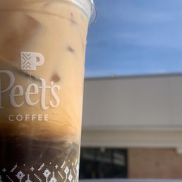 Coconut Black Tie - A clear plastic cup filled with iced coffee from Peet's Coffee, displaying the Peet's logo on the front. The cup contains ice cubes and a light brown coffee beverage. The background shows a blue sky with some clouds and part of a building, indicating an outdoor setting.