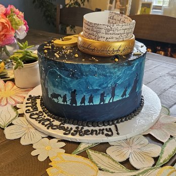 The Yummery - A round birthday cake features a silhouette scene of people walking against a blue sky, decorated with gold accents and topped with ribbons and gold beads. - Cupcakes Near Me - White Marsh, Maryland