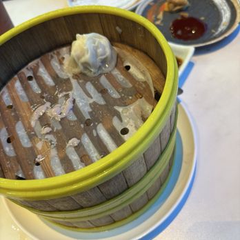 Juicy Dumplings - Pork dumpling - A steamed dumpling is positioned inside a bamboo steamer basket, which has a yellow rim and a perforated base. The basket contains some leftover filling and sauce residue. It is placed on a white plate, with other plates and condiments visible on the table, indicating a dining setting at a restaurant that serves dim sum or similar cuisine. - Chinese Near Me - Euless, Texas
