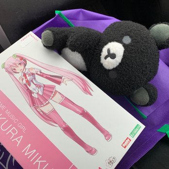 AnimeStuffStore.com - Hatsune Miku Figure, and Rilakkuma Plush - The image shows a black plush bear with gray ears and a white nose, positioned on a purple bag inside a vehicle. Next to the plush toy is a box featuring an anime character with long pink hair, dressed in a pink and white outfit, labeled 'Sakura Miku.' The scene appears to be inside a car, with part of the vehicle's interior visible. - Toy Stores Near Me - Plainfield, Illinois