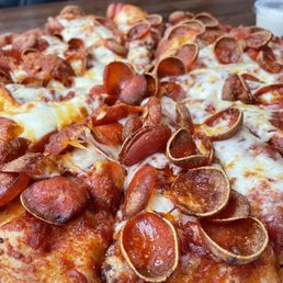 Pepperoni Pizza - A close-up image of a pepperoni pizza featuring a golden-brown crust, melted cheese, and numerous slices of pepperoni. The pizza is placed on a table, with a small container of dipping sauce visible in the background. The focus is on the toppings and cheese, highlighting the texture and appearance of the pizza.