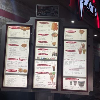 Oscar's Frozen Custard - Drive thru menu and ordering speaker - Outdoor illuminated menu board at Oscar's Frozen Custard features sections for burgers, chicken, salads, soups, drinks, sundaes, and frozen custard. The menu includes items like Oscar Burgers, chicken sandwiches, specialty sundaes, and various frozen custard sizes. The board is positioned on a gravel surface with a brick wall and a business sign visible in the background. - Burgers Near Me - Franklin, Wisconsin