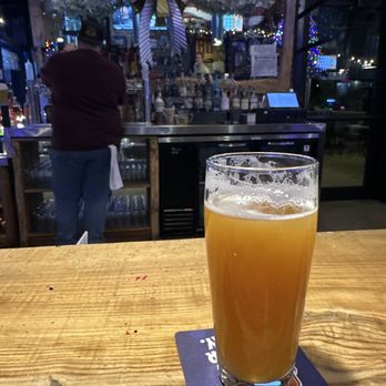 Ohio Brewing - A tall, transparent glass filled with beer featuring a foamy head is positioned on a coaster on a wooden table. The background shows the interior of Ohio Brewing, including a bar area with bottles, a bartender, and festive decorations such as garlands and a large ribbon bow. The setting appears to be a casual brewery and restaurant environment. - Breweries Near Me - Columbus, Ohio