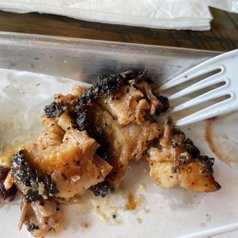 Duke City BBQ - A partially eaten serving of barbecue ribs with blackened, charred edges and visible meat, placed on a tray lined with paper. A white plastic fork rests on the tray, and a small cup of barbecue sauce is visible on the right side of the image. The background includes a wooden table surface and a napkin. - Barbeque Near Me - Albuquerque, New Mexico