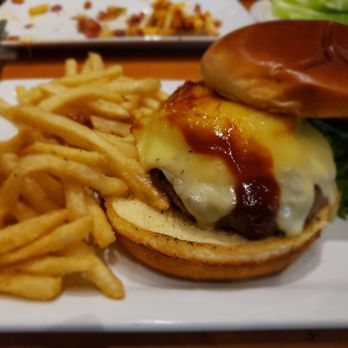 Treehouse Pub & Eatery - Hawaiian burger - A cheeseburger with a beef patty, melted cheese, a fried egg, and barbecue sauce is presented on a toasted bun with lettuce. French fries are served on the side, and the burger is placed on a white rectangular plate. The background shows a blurred view of a restaurant setting with additional plates and utensils. - Pizza Near Me - Bettendorf, Iowa