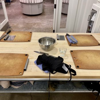 Sur La Table - Sur La Table cooking class - A cooking class setup features four wooden cutting boards with knives and utensils placed on each. A metal mixing bowl, a glass of water, and cloth towels are also on the table. The table is situated in a room with a visible doorway leading to a kitchen or retail space, and a fire extinguisher is mounted on the wall. The setup appears prepared for a cooking demonstration or class. - Kitchen Supplies Near Me - Glendale, Wisconsin