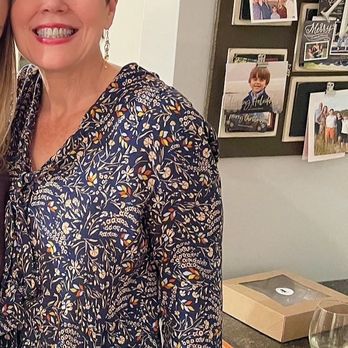 Covet - A woman with short dark hair, wearing a long-sleeved dress with a floral pattern, is smiling at the camera. She is standing indoors near a wall decorated with a collage of photographs and a small sign. The background includes a countertop with a wine glass and a salad, and a cabinet below. The setting appears to be a casual gathering or celebration at home. - Women's Clothing Near Me - Boston, Massachusetts