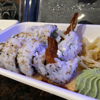 jony's sushi - Shrimp tempura roll - A sushi roll featuring shrimp tempura and sesame seeds is arranged in a white takeout container. Accompanying the roll are slices of pickled ginger, a dollop of wasabi, and a small amount of sauce. The background shows a wet window and part of a sushi restaurant setting, indicating a takeout or dine-in meal. - Sushi Bars Near Me - Columbus, Ohio