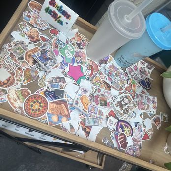 The Rabbit Hole - A wooden table holds an assortment of stickers, two drinks with straws, and potted plants, with a sign that says 'Stickers & Stuff.' - Tea Rooms Near Me - Eugene, Oregon