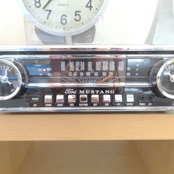 The Sound Guy - Ion record player - A vintage Ford Mustang car radio with a chrome finish, featuring two large control knobs for volume and tuning, a central display for radio frequencies, and several buttons for functions like source, back, and pause. The radio is positioned on a light-colored wooden surface, with a wall clock visible in the background. The radio has a classic design with branding and control labels. - Electronics Repair Near Me - Phoenix, Arizona