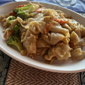 Pha Khao - Chicken pad see ew - A dish of chicken pad see ew with broccoli is served on a white oval plate, accompanied by drinks on a dining table. - Thai Near Me - Kenosha, Wisconsin