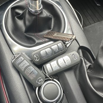 Quick Car Keys - OEM keys. I bought 2. - The photo depicts the interior of a vehicle, focusing on the center console area. There is a gear shift with a black leather cover and red stitching, three Mazda key fobs with buttons for lock, unlock, and trunk release, and a keychain attached to one of the fobs. The console also features a circular control knob and additional buttons, with a charging cable visible in the background. - Keys & Locksmiths Near Me