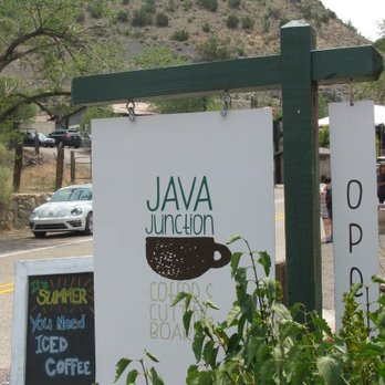 Java Junction - The image shows a sign for Java Junction coffee shop, featuring a stylized coffee cup graphic and text. The sign is mounted on a green post, with a mountain and parked cars visible behind it. There is also a smaller chalkboard sign advertising iced coffee, positioned on the sidewalk. - Bed & Breakfast Near Me - Madrid, New Mexico