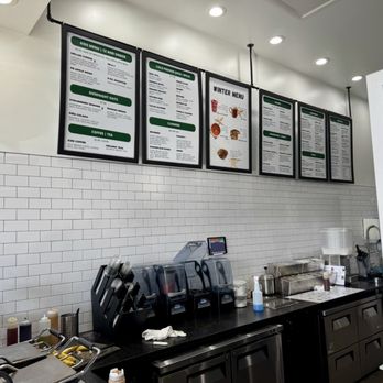 Extract Juicery - Four menu boards are suspended from the ceiling above a black countertop in a juice bar or cafe. The boards display various food and drink options, including kids' menus, cold pressed juices, and winter specials. The counter below has equipment, bottles, and utensils, with a white subway tile wall behind. A small digital device is also visible on the counter. - Juice Bars & Smoothies Near Me - St. Charles, Illinois