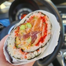 Sushi Burritos - A sushi burrito containing ingredients like fish, avocado, and hot Cheetos is held in front of a car's steering wheel, showcasing its cross-section.