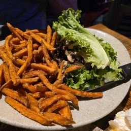 Bison Burger - A ceramic plate holds a serving of crispy, golden-brown sweet potato fries alongside a side salad consisting of green leaf lettuce and sliced mushrooms. The plate is placed on a wooden table, with a knife resting on the edge of the salad. The background shows a dimly lit dining environment.