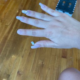 the broken pointer finger nail. - A hand with long, acrylic nails featuring decorative designs is displayed over a wooden floor, with a small black stool in the background.