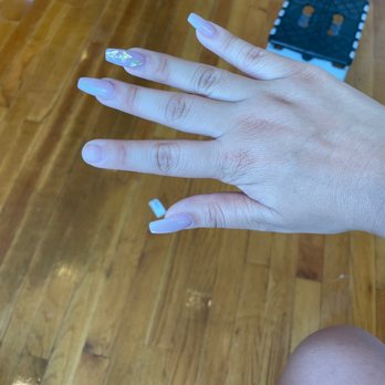 Kiwi Nail - the broken pointer finger nail. - A hand with long, acrylic nails featuring decorative designs is displayed over a wooden floor, with a small black stool in the background. - Nail Salons Near Me - Harrison, New York