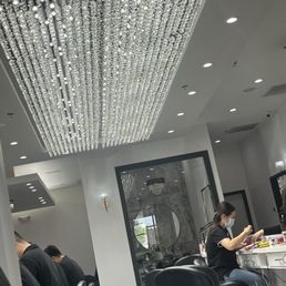 A nail salon interior featuring a large, ornate crystal chandelier, clients seated in black chairs, and a technician working on a client while wearing a face mask.