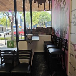 Seating - The image shows an interior seating space at Monster Donuts, with black chairs arranged along the wall and near a window. The window provides a view of trees, parked cars, and a street scene outside. The ceiling has a wooden finish, and there is a decorative black drip design on the window frame. The pink wall on the right side displays donut-themed artwork, including images of donuts and the Monster Donuts logo. The floor appears to be dark, possibly marble or tile, and the space is well-lit by natural light from the window.