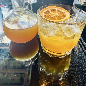 Honey Elixir Bar - Sunshine of your love | calendula, chamomile, turmeric, pink peppercorn, yerba maté, orange - A citrus cocktail with ice and a dried orange slice is placed next to a decanter containing a similar drink, on a reflective dark surface. - Lounges Near Me - Denver, Colorado