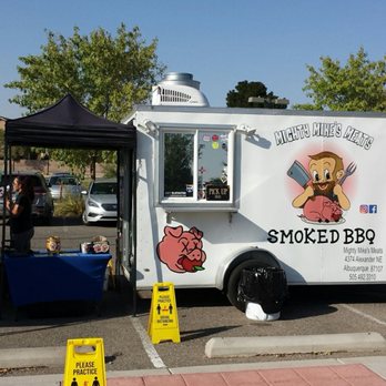 Mighty Mike's Meats - Home for happy carnivores. - A white food truck called Mighty Mike's Meats offering smoked BBQ, with cartoon graphics, parked in a lot with people and signs. - Food Trucks Near Me - Albuquerque, New Mexico
