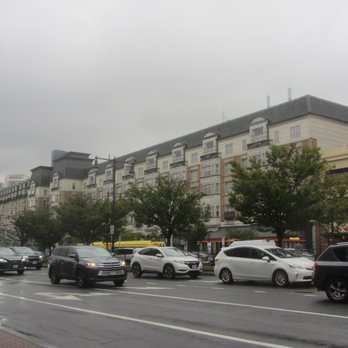 Kenmore Square - The image shows a city street with multiple vehicles, including sedans, SUVs, and a bus, moving along the road. A row of multi-story buildings with retail storefronts and residential units is visible in the background, with trees lining the sidewalk. The sky is overcast, and the scene appears to be in an urban commercial area. - Landmarks & Historical Buildings Near Me - Boston, Massachusetts