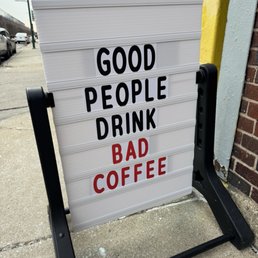 A sidewalk sign with a message reading 'Good People Drink Bad Coffee' in black and red letters, placed outside a business.