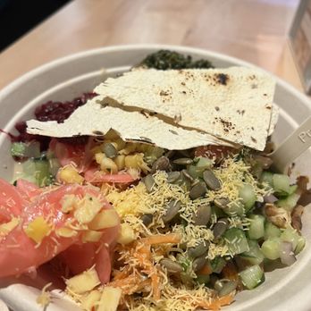 RASA - A takeout bowl holds a salad with chopped cucumbers, shredded cheese, sunflower seeds, and pieces of flatbread on top. The salad includes various vegetables and toppings, presented in a disposable container on a wooden table. - Indian Near Me - Fairfax, Virginia