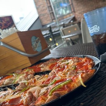 Pace's Pizzeria - The Pace's Special. - A pizza with assorted toppings such as green peppers, onions, and meat is placed on a black tray on a table. The background shows an outdoor dining area with brick walls, windows, and a grill, indicating a restaurant patio setting. A glass of soda and a cardboard container are also visible on the table. - Pizza Near Me - Jamestown, New York