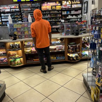 RaceTrac - a man in an orange hoodie in a store - A person dressed in an orange hoodie and dark pants is standing at a checkout counter in a convenience store. The counter displays various snacks and candies, with shelves of cigarettes and tobacco products behind. Digital screens are visible above the counter, and a cashier is attending to the customer. The store has tiled flooring and various products on display, including snacks, cigarettes, and small items on racks. - Gas Stations Near Me - St Augustine, Florida