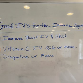 Micronutrient Infusion Services - A whiteboard features handwritten text in blue ink listing IV treatments for immune system support, including 'Immune Boost IV & Shot,' 'Vitamin C IV 20G or more,' and 'Dragonfire or Myers.' Three colored magnets (blue, orange, black) are attached next to each line of text, possibly indicating different options or categories. The background shows reflections of ceiling lights, and the overall setting appears to be an indoor space, likely a clinic or health center. - Oxygen Bars Near Me - Albuquerque, New Mexico