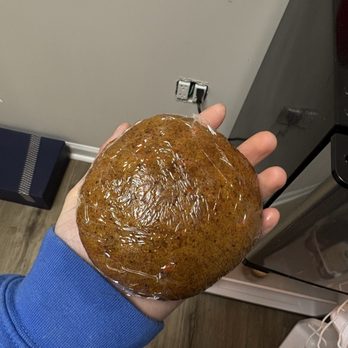 Yorgo's Bageldashery - Carrot cake cookie (more like a whoopie pie) - A person in a blue sleeve holds a large, round baked item covered with a shiny glaze, in a kitchen with a gray wall and appliances. - Bagels Near Me - Norfolk, Virginia
