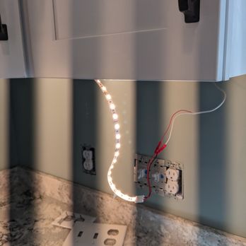 Daigle Electric - Mike Daigle suggested these lights instead of the big box ones I was thinking about - I'm glad I listened to him. - A strip of LED lights is attached to a wall above a kitchen counter, connected to an electrical outlet with red and white wires. The outlet cover is removed, exposing the wiring. The LED strip is illuminated, casting light on the wall and counter surface. The scene appears to be part of an electrical or lighting installation process. - Electricians Near Me - Barrington, New Hampshire