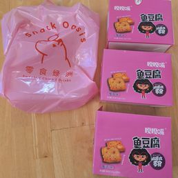 Three pink boxes of fish tofu snacks with cartoon character illustrations and a pink plastic bag from Snack Oasis are placed on a wooden surface.