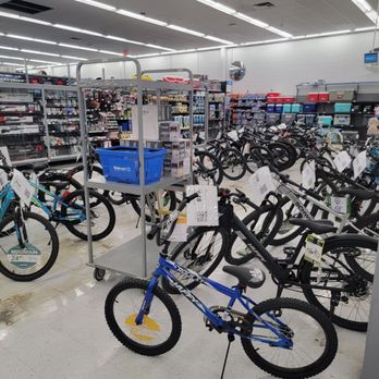 Walmart Supercenter - Bikes - Multiple bicycles are displayed inside a Walmart store, arranged in rows with price tags attached. A blue mountain bike is prominently positioned in the foreground, with other bikes of different styles and sizes visible behind it. The store has bright overhead lighting, and shelves stocked with various products are visible in the background. A metal cart with a blue Walmart shopping basket is also present near the bikes. - Department Stores Near Me - Willows, California