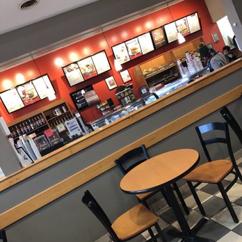 Big Apple Bagels - Menu and ordering - A fast-food restaurant interior featuring a red accent wall with illuminated menu boards, a counter with food display, and seating with wooden tables and black chairs. - Bagels Near Me - Davison, Michigan