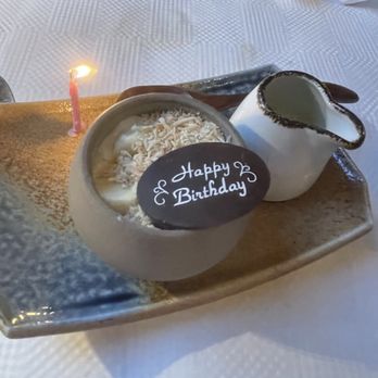 Le Colonial - Vietnamese pudding - A birthday dessert featuring a small bowl with a "Happy Birthday" chocolate sign, a lit candle, and a small pitcher, all on a ceramic plate. - Vietnamese Near Me - Chicago, Illinois