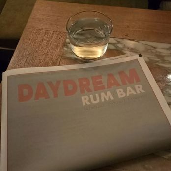 Daydream - A clear glass of water is positioned on a wooden table with a mixed wood and marble surface. A menu from Daydream Rum Bar, featuring a pastel-colored design with large text, is partially visible in the foreground. The setting appears to be a dimly lit indoor space, likely a bar or restaurant. - Cocktail Bars Near Me - Albuquerque, New Mexico