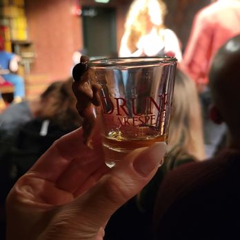 Drunk Shakespeare - Chicago - souvenir shot glass - A person holds a shot glass with a small figurine of a woman attached, in a dimly lit setting with people and stage performers in the background. - Performing Arts Near Me - Chicago, Illinois