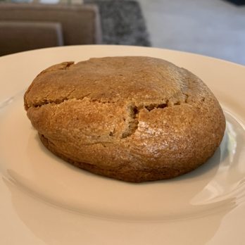 ThoroughBread - Banana Nutella cookie - GET THIS NOW - A large, round cookie with a cracked, golden-brown surface is placed on a plain white plate. The cookie appears thick and soft, with a slightly uneven texture and a visible crack on the top. The background shows a blurred indoor setting, possibly a bakery or cafe environment. - Bakeries Near Me - Austin, Texas