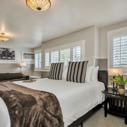 Craftsman Inn - King bed with white bedsheets, brown throw blanket, and two striped pillows. Side table and lamp. Shutters. Sofa bed. Wall art and wall lamp