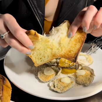 Platea - A person is tearing a piece of bread over a plate of oysters topped with sauce and lemon slices, at a restaurant setting. - Peruvian Near Me - Pinecrest, Florida