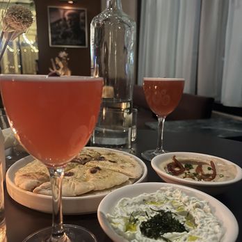 Nerina - The Loukoumi Cocktail - The image shows a dining table with two glasses of pinkish drinks, a clear glass water bottle, and three bowls containing dips and flatbread. The background includes a wooden cabinet with decorative items, a framed picture, and curtains, indicating an indoor restaurant setting. The table setup suggests a Mediterranean or Greek-inspired meal. - Greek Near Me - Brooklyn, New York