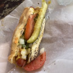 Hot Char Jumbo Dog - A hot dog features a sausage in a bun topped with mustard, a pickle spear, slices of tomato, and chopped onions. The hot dog is served on crumpled parchment paper, with a rustic wooden surface partially visible in the background.