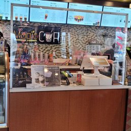 the inside of a coffee shop - The image shows a coffee shop counter with digital menu boards displaying drink options and prices. There is a glass display case on the left containing baked goods, and promotional signs and flyers are placed on the counter. The background includes a mosaic tile wall and additional menu screens, indicating a contemporary coffee shop environment.