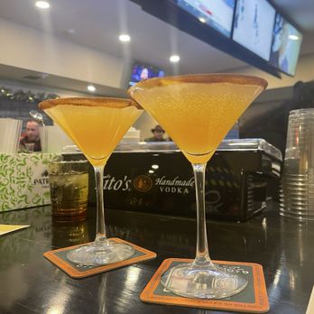 Ljs Kitchen - Apple cider martini - Two apple cider martinis in cocktail glasses are placed on a bar counter, with a background of TV screens and bar items. - Pizza Near Me - Woonsocket, Rhode Island