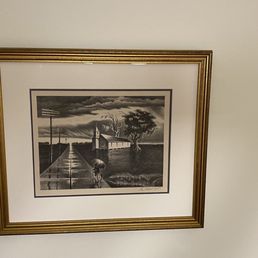 Hand signed original artwork, double matted, under museum glass. Sorry for the bad lighting. - A black-and-white artwork in a gold-colored frame is mounted on a plain, light-colored wall. The artwork depicts a rural scene with a road, a bicycle, a small house, trees, and a cloudy sky. The frame has a white mat border that surrounds the image, and the wall is unadorned.